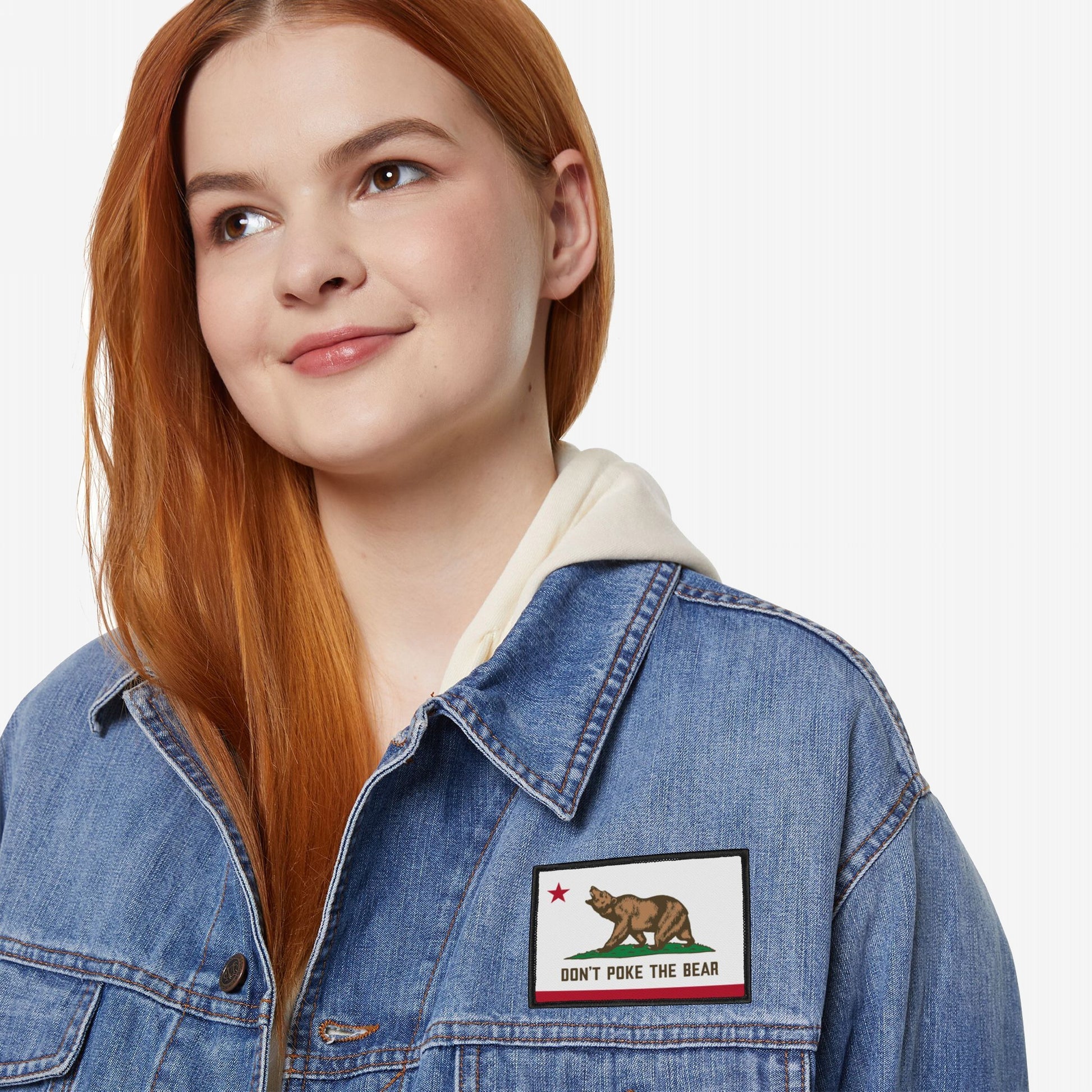 A young person with red-brown hair wears a denim jacket featuring the California Don't Poke The Bear Flag Patch, showcasing a bear and star with a warning message.
