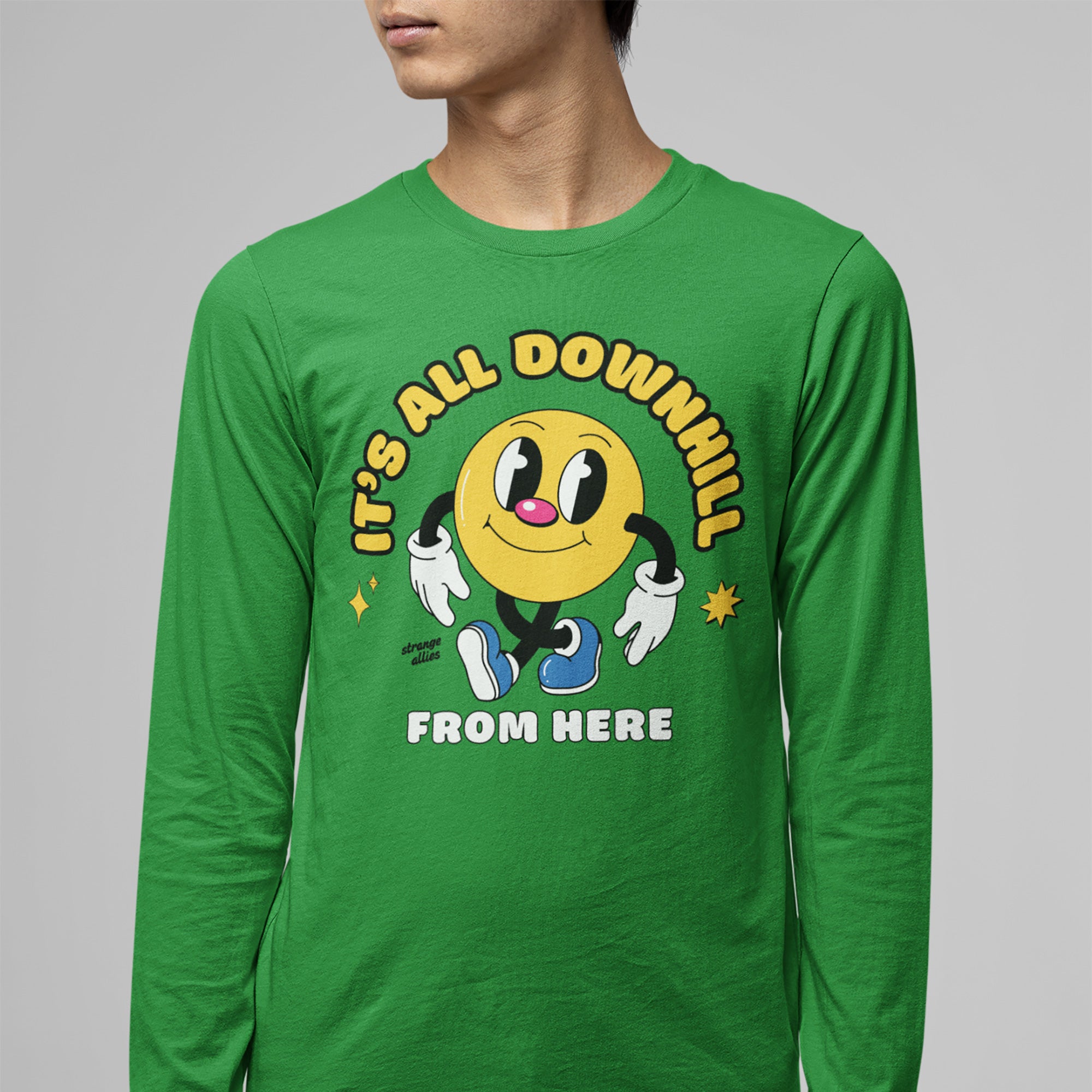 A person wears the Downhill Shirt showcasing a smiley-face character with text IT’S ALL DOWNHILL FROM HERE, embodying playful cynicism. A strange allies signature and star accents complete the design.