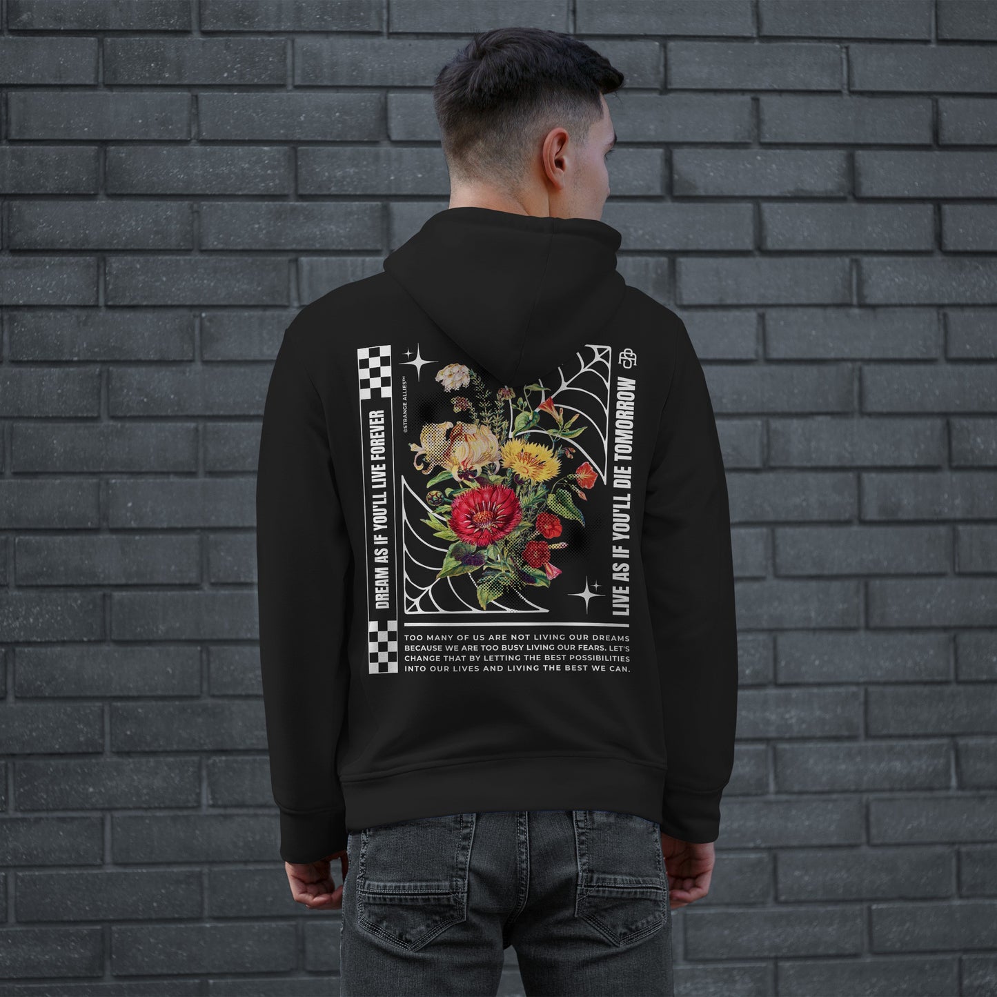 Man wearing the Dreams Hoodie, featuring a colorful floral design within a spider-web frame, checkerboard accents, and motivational text, embodying defiance and ambition against a gray brick wall.