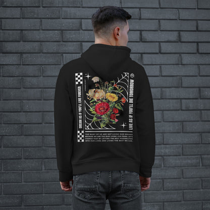 Man wearing the Dreams Hoodie, featuring a colorful floral design within a spider-web frame, checkerboard accents, and motivational text, embodying defiance and ambition against a gray brick wall.