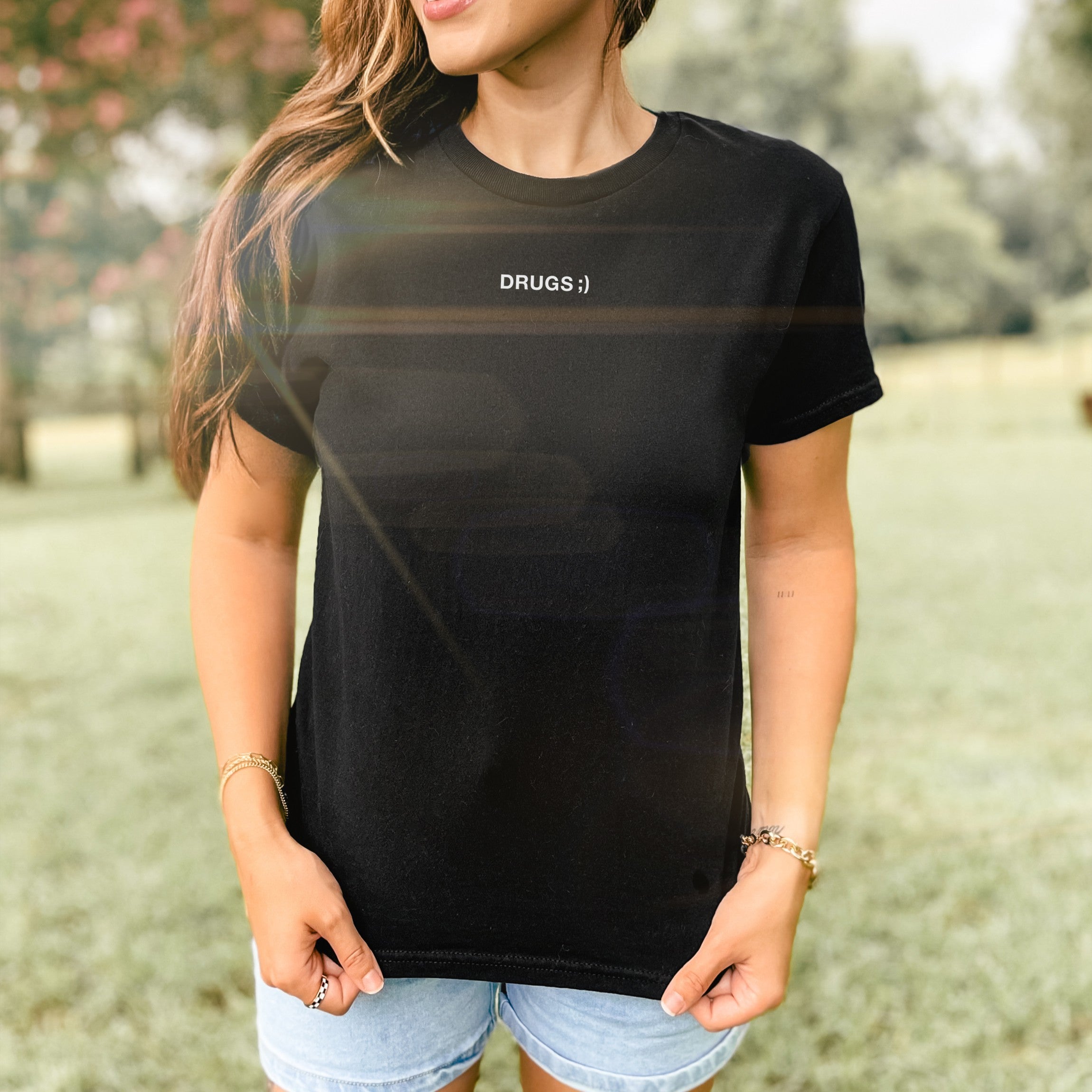Person outdoors in “DRUGS ;)” black tee, light denim shorts. The Drugs Women's Baby Tee embodies minimalist chaos, ideal for casual outings, aligning with bold, carefree expressions.