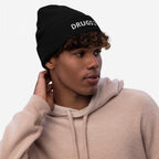 Young man in a black knit beanie with DRUGS; ) embroidered, wearing a light-tan hoodie, looking right with hand on neck, embodying a bold, ironic statement from the Drugs Hat collection.