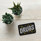 Drugs Patch: A rectangular black patch embroidered with “DRUGS” in bold white letters, accented by yellow starburst motifs, on a light-wood tabletop alongside two potted succulents in white ceramic pots.