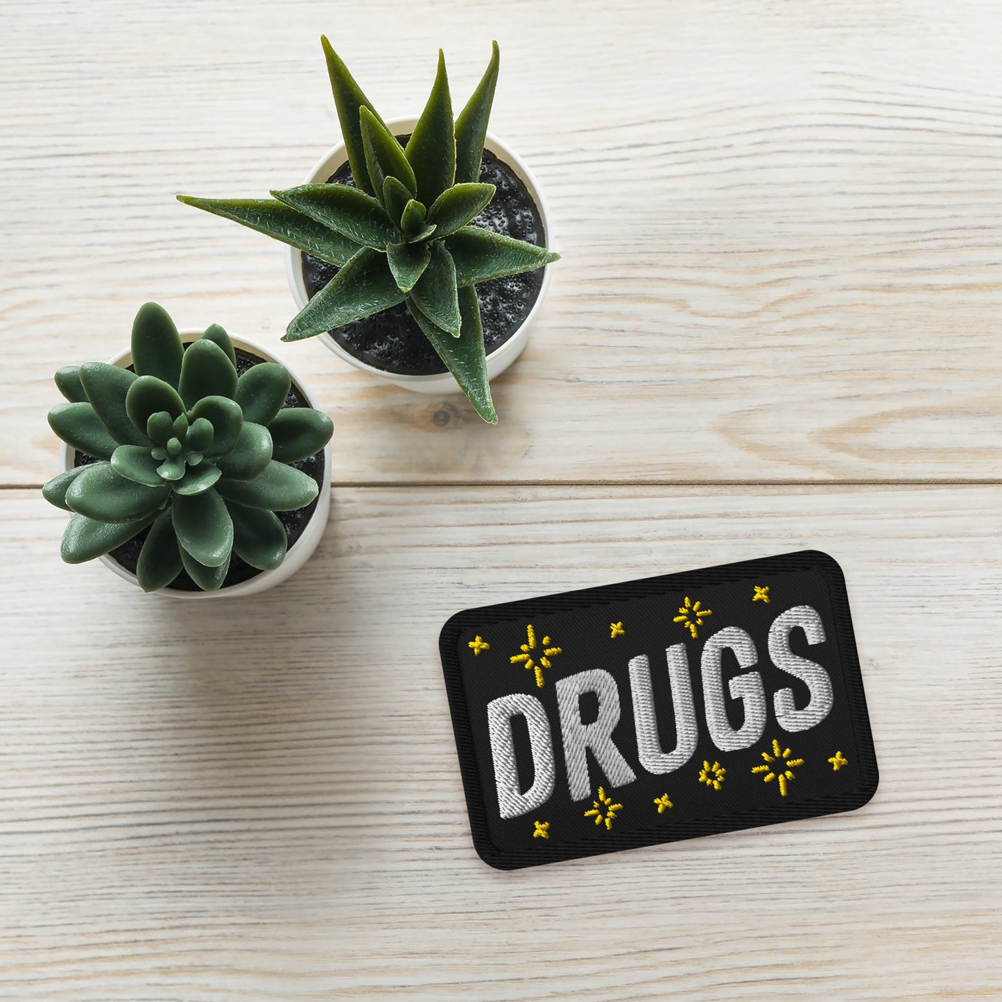 Drugs Patch: A rectangular black patch embroidered with “DRUGS” in bold white letters, accented by yellow starburst motifs, on a light-wood tabletop alongside two potted succulents in white ceramic pots.