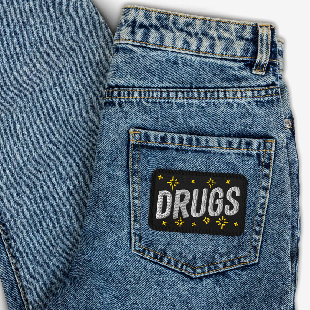 Close-up of the Drugs Patch, featuring bold white lettering and yellow star motifs, embroidered on a back pocket of blue denim jeans, reflecting a bold, rebellious spirit.