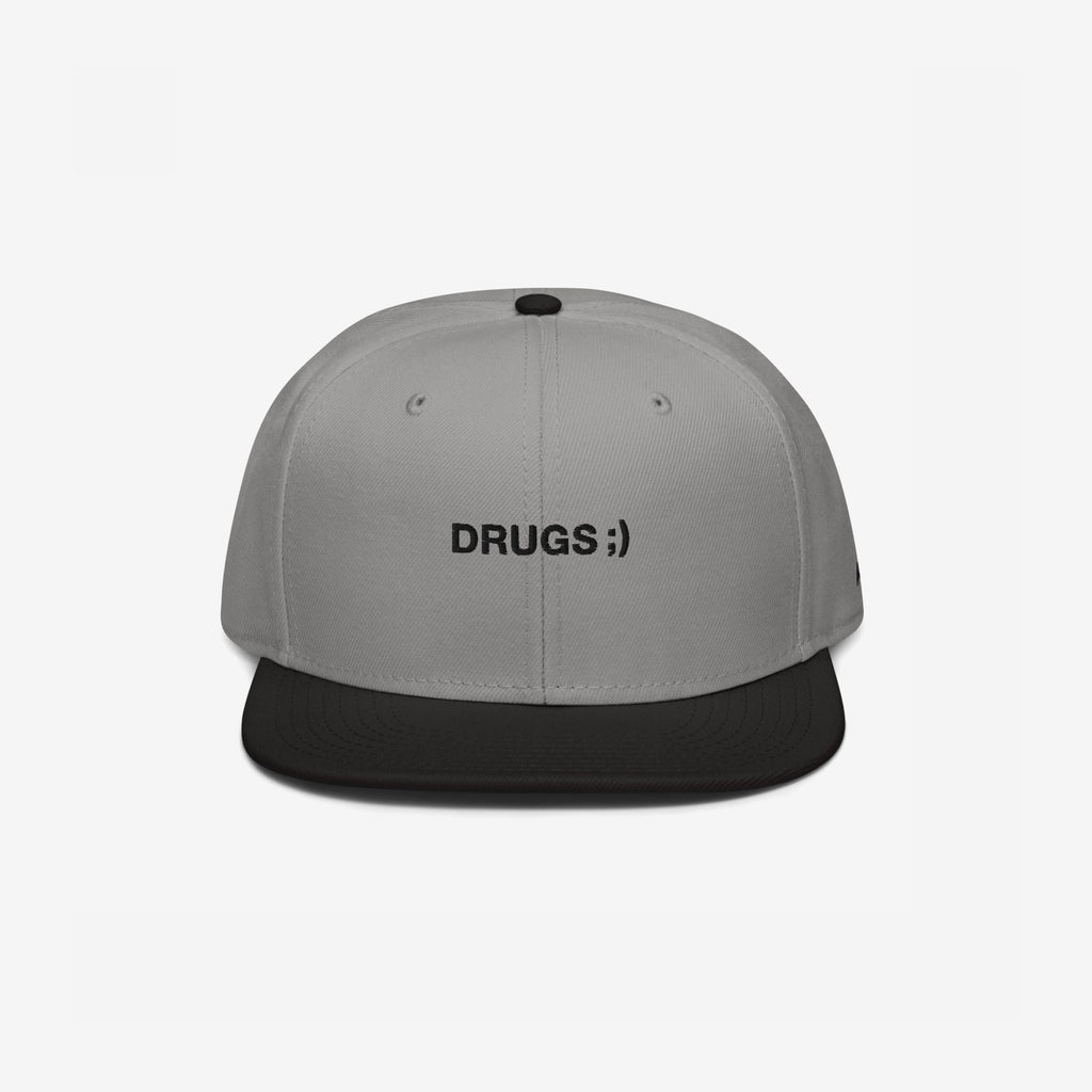 Two-tone snapback cap titled Drugs Hat with a light gray crown and black brim, featuring DRUGS ;) embroidered on the front, encapsulating a bold, ironic statement.