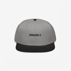 Two-tone snapback cap titled Drugs Hat with a light gray crown and black brim, featuring DRUGS ;) embroidered on the front, encapsulating a bold, ironic statement.
