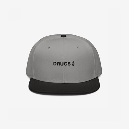 Two-tone snapback cap titled Drugs Hat with a light gray crown and black brim, featuring DRUGS ;) embroidered on the front, encapsulating a bold, ironic statement.