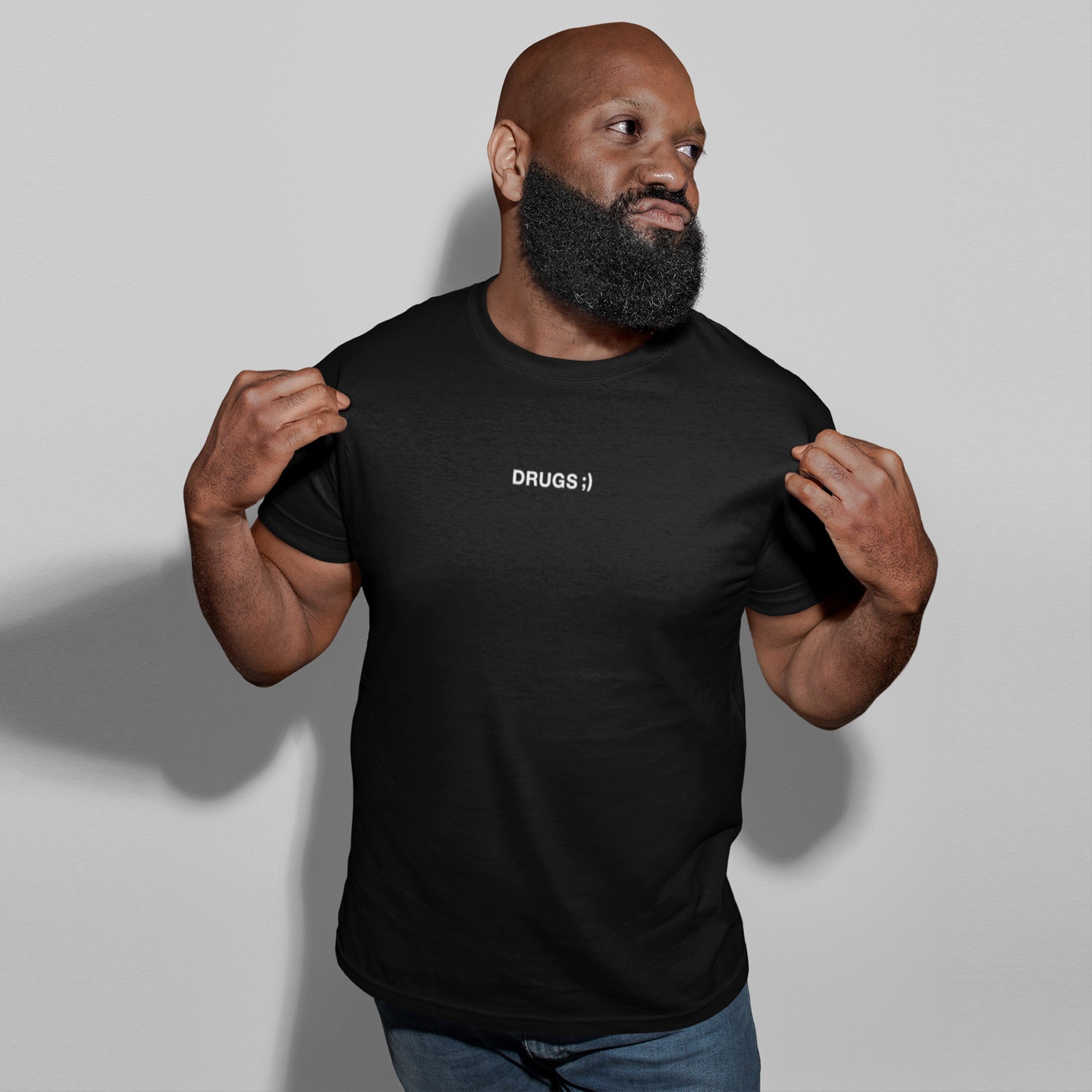 Muscular man wearing a black Drugs T-shirt with ironic text, pulling the shirt to emphasize its bold design, capturing the unapologetic and minimalist style that defines the product's message.
