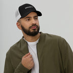 Bearded man in olive bomber jacket wearing a DRUGS :) trucker hat, showcasing its bold, ironic embroidery. The cap adds edgy flair, aligning with a playful, statement-making style.