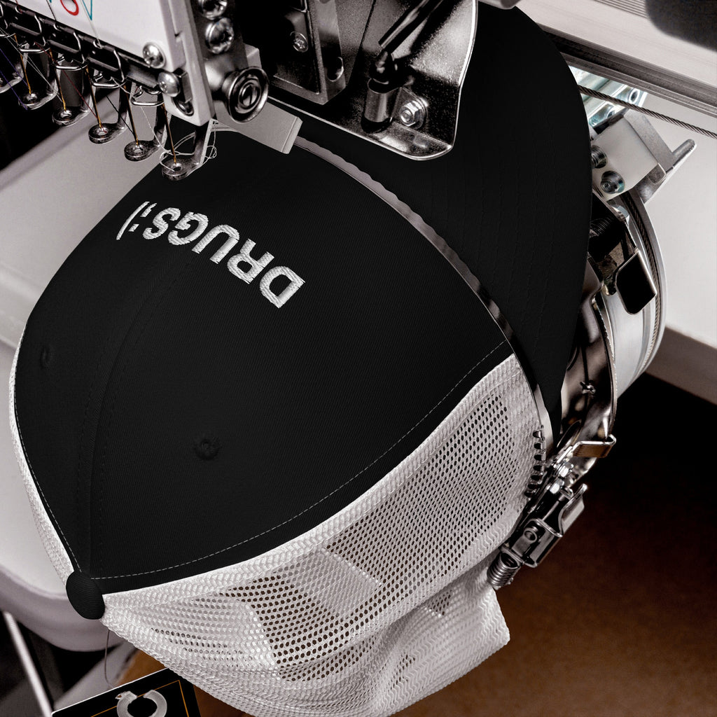 Industrial embroidery machine stitching the word “DRUGS!” on a black-and-white mesh trucker cap, capturing the bold, ironic vibe of the Drugs Hat.