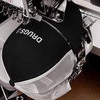 Industrial embroidery machine stitching the word “DRUGS!” on a black-and-white mesh trucker cap, capturing the bold, ironic vibe of the Drugs Hat.