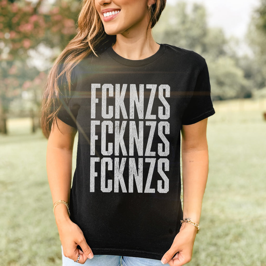 Person outdoors in a park wearing the FCKNZS Women's Baby Tee, featuring distressed FCKNZS text stacked thrice, symbolizing anti-fascist sentiment. The wearer holds the shirt's hem and smiles confidently.