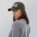A woman wears a FCKNZS Hat, a green and brown camouflage baseball cap with KNZS embroidered in white on the front, symbolizing anti-fascist sentiment. She’s turned slightly over her shoulder.