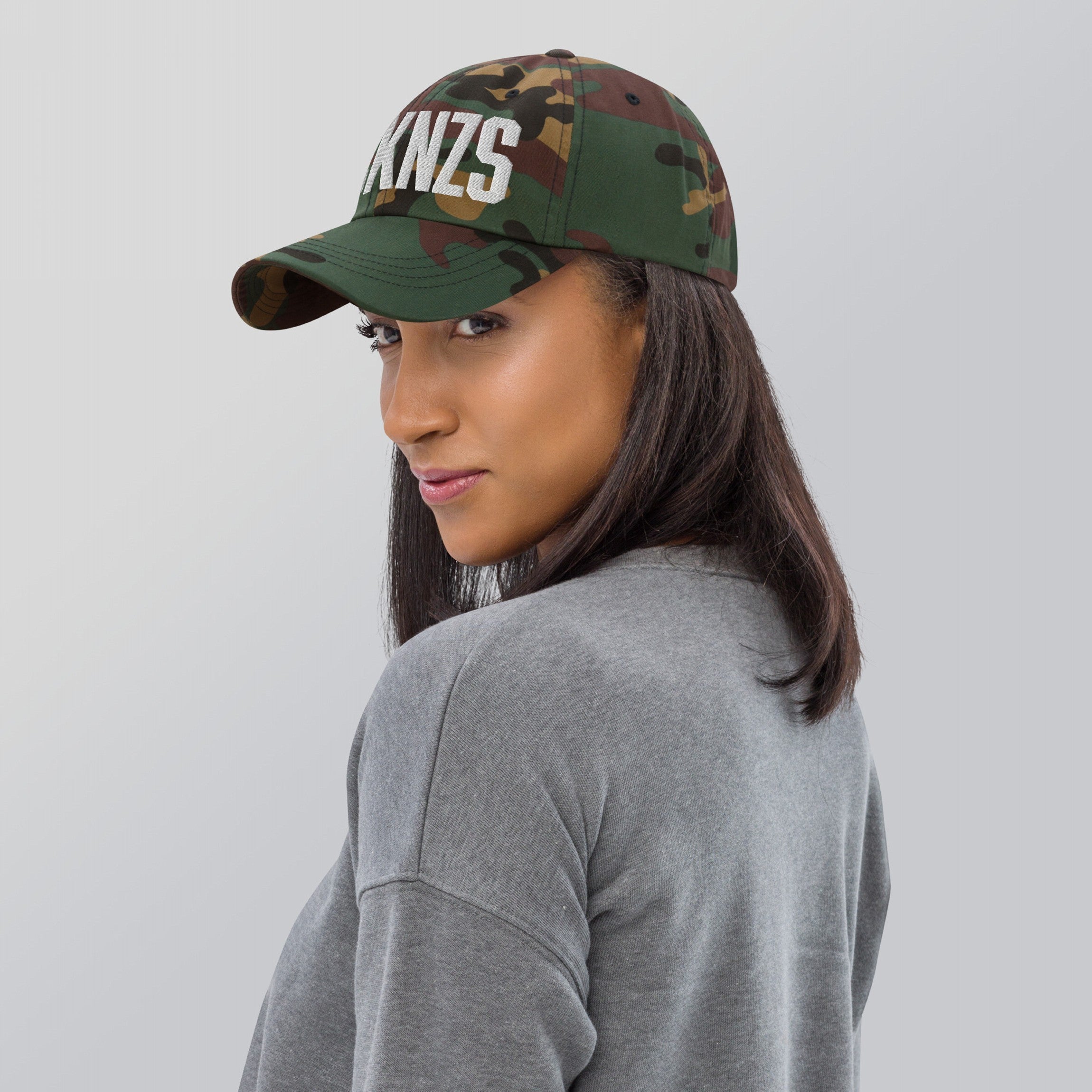 A woman wears a FCKNZS Hat, a green and brown camouflage baseball cap with KNZS embroidered in white on the front, symbolizing anti-fascist sentiment. She’s turned slightly over her shoulder.