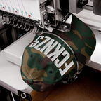 Camouflage-print baseball cap clamped into embroidery machine, with FCKNZS being stitched in bold white letters across the front, showcasing an anti-fascist protest message.