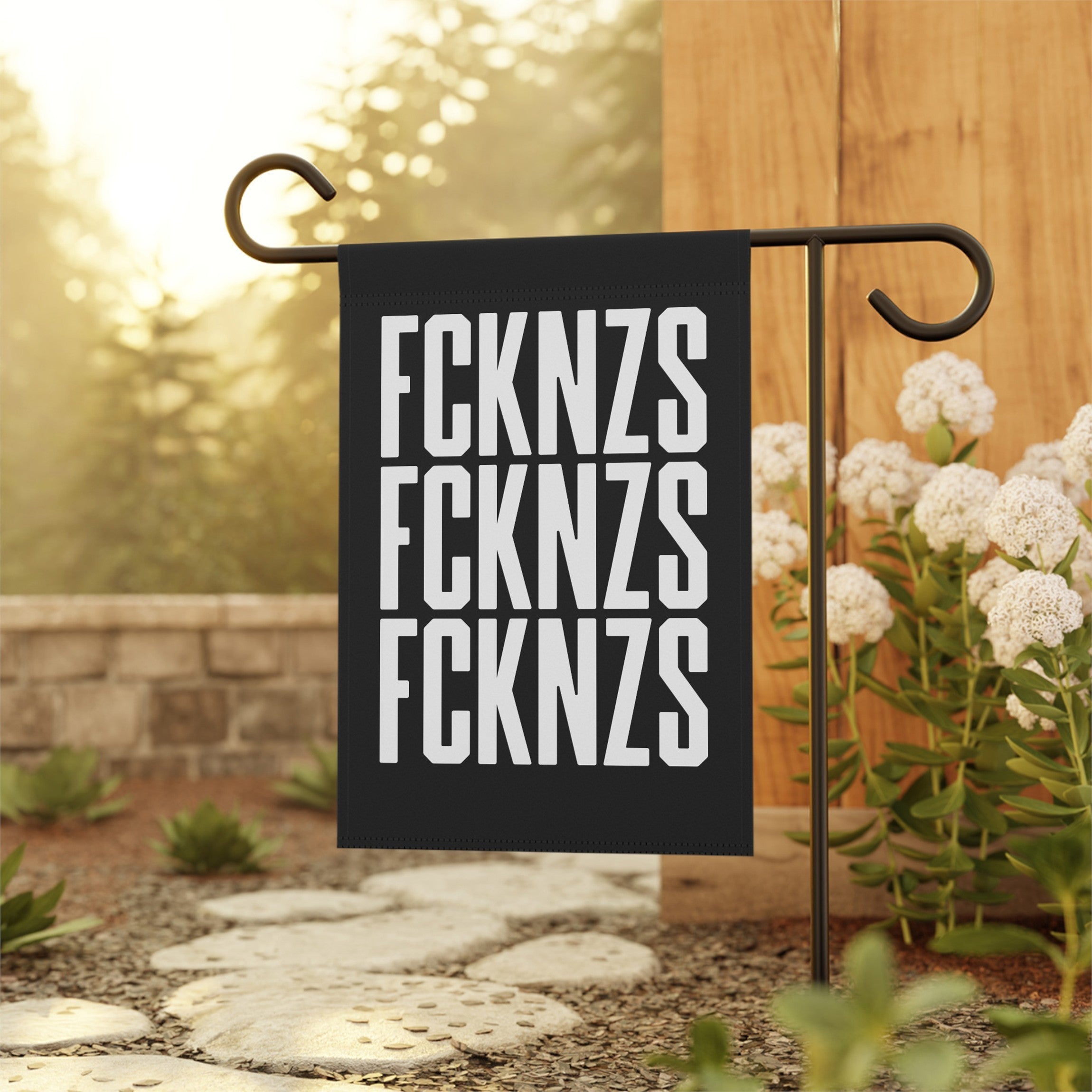 FCKNZS Garden Flag displayed in a garden, featuring bold white letters on a black background. Mounted on a metal stand beside a stone path and flowers, conveying a clear anti-fascist message.