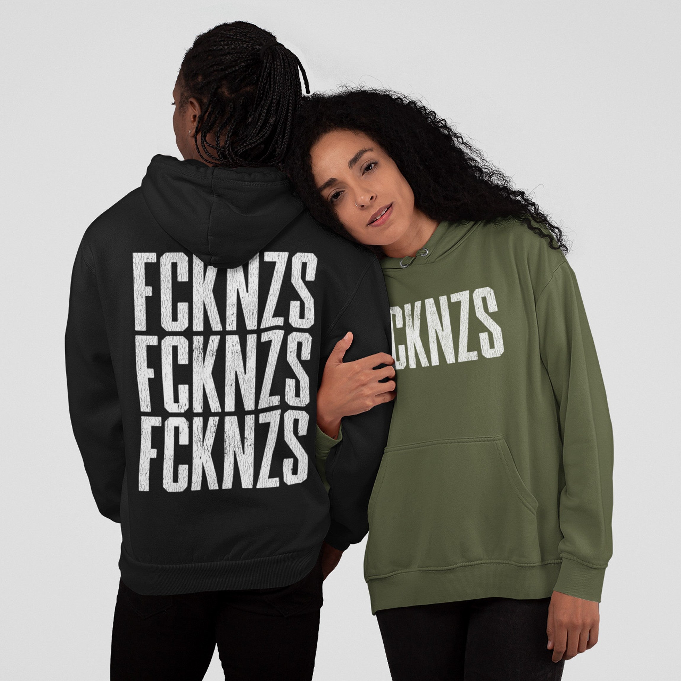 Two people wearing FCKNZS Hoodies; one black with bold back print, the other olive-green with front print. The design promotes anti-fascism and equality, reflecting strong social values.