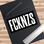 FCKNZS Sticker & Magnet on a black mat with bold white lettering, accompanied by writing tools and a ruler. An anti-fascist design emphasizing clear visibility and strong messaging against hate.