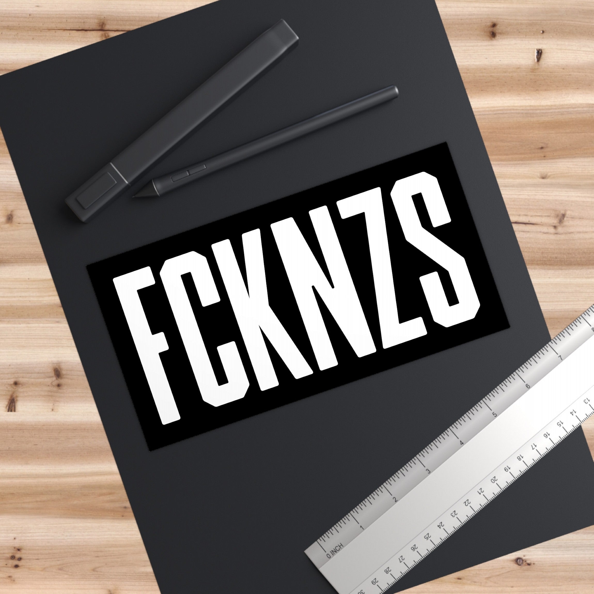 FCKNZS Sticker & Magnet on a black mat with bold white lettering, accompanied by writing tools and a ruler. An anti-fascist design emphasizing clear visibility and strong messaging against hate.