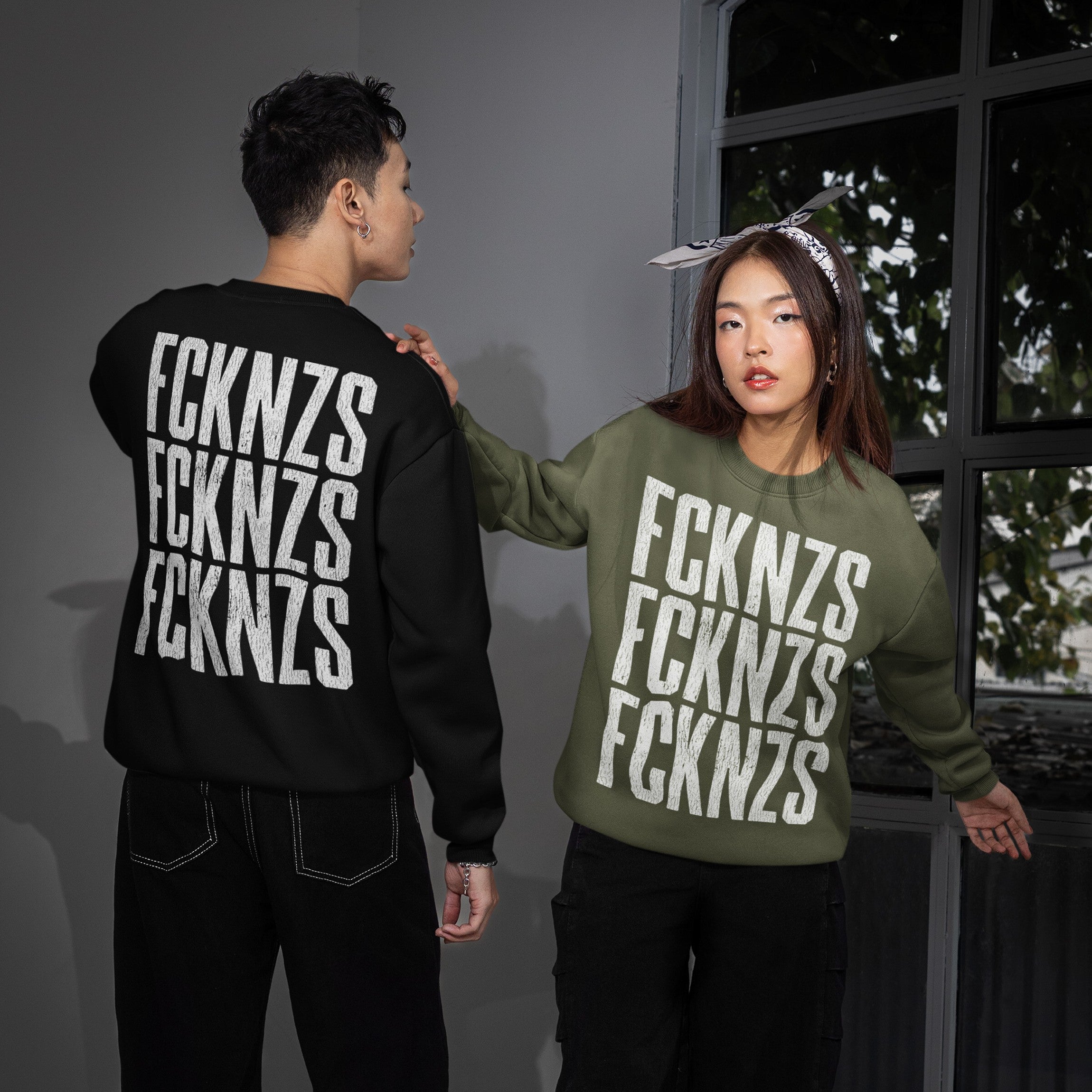 Models showcase the FCKNZS Sweatshirt, featuring bold anti-fascist text. One wears a black version viewed from behind, the other an olive green from the front, embodying protest spirit.