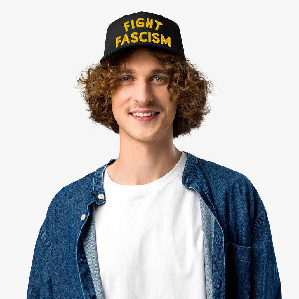 A young person smiles while wearing the Fight Fascism Hat, featuring bold yellow FIGHT FASCISM lettering on a black cap, paired with a denim shirt and white T-shirt.