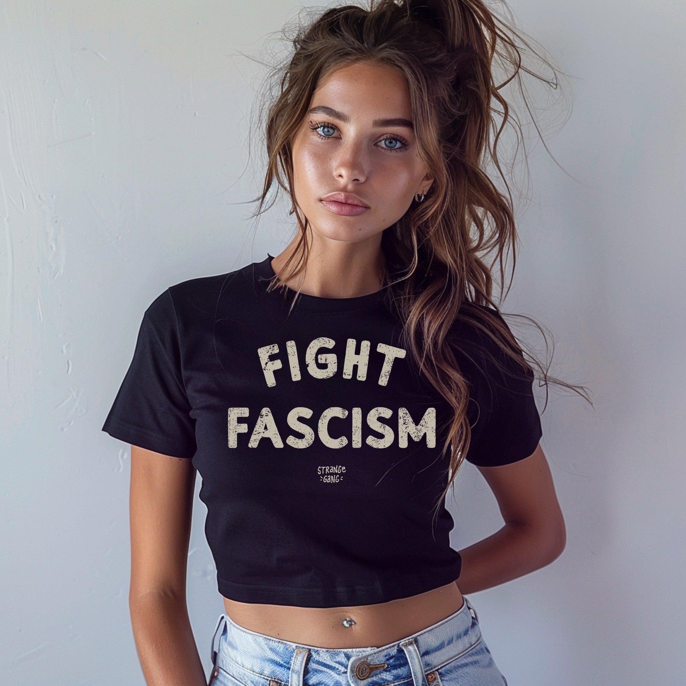 Young woman models the Fight Fascism Women's Baby Tee, featuring bold cream lettering on a black cropped T-shirt, paired with light-wash jeans, embodying a message of defiance and equality.