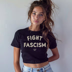 Young woman models the Fight Fascism Women's Baby Tee, featuring bold cream lettering on a black cropped T-shirt, paired with light-wash jeans, embodying a message of defiance and equality.