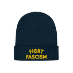 The Fight Fascism Hat is a dark-blue ribbed knit beanie with a folded cuff, featuring bold yellow FIGHT FASCISM embroidery, i