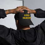 Person wearing the Fight Fascism Hat, showcasing yellow FIGHT FASCISM text on a black cap, symbolizing anti-fascist stance; tattoos and hoop earring visible, embodying bold activism and equality.