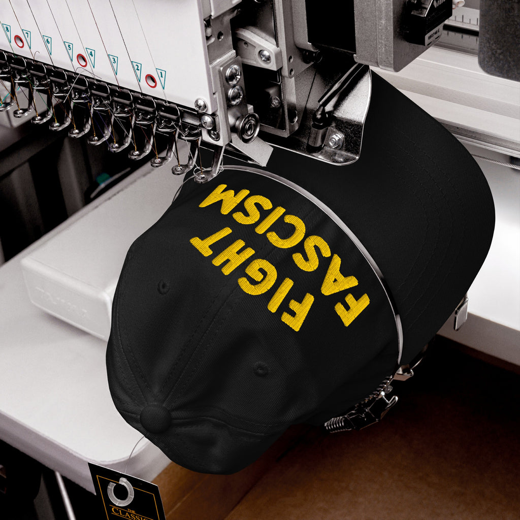 Fight Fascism Hat in embroidery machine, yellow thread stitching bold slogan on black cap, showcasing anti-fascist message for visibility in protests, marches, and daily life.