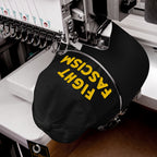 Fight Fascism Hat in embroidery machine, yellow thread stitching bold slogan on black cap, showcasing anti-fascist message for visibility in protests, marches, and daily life.
