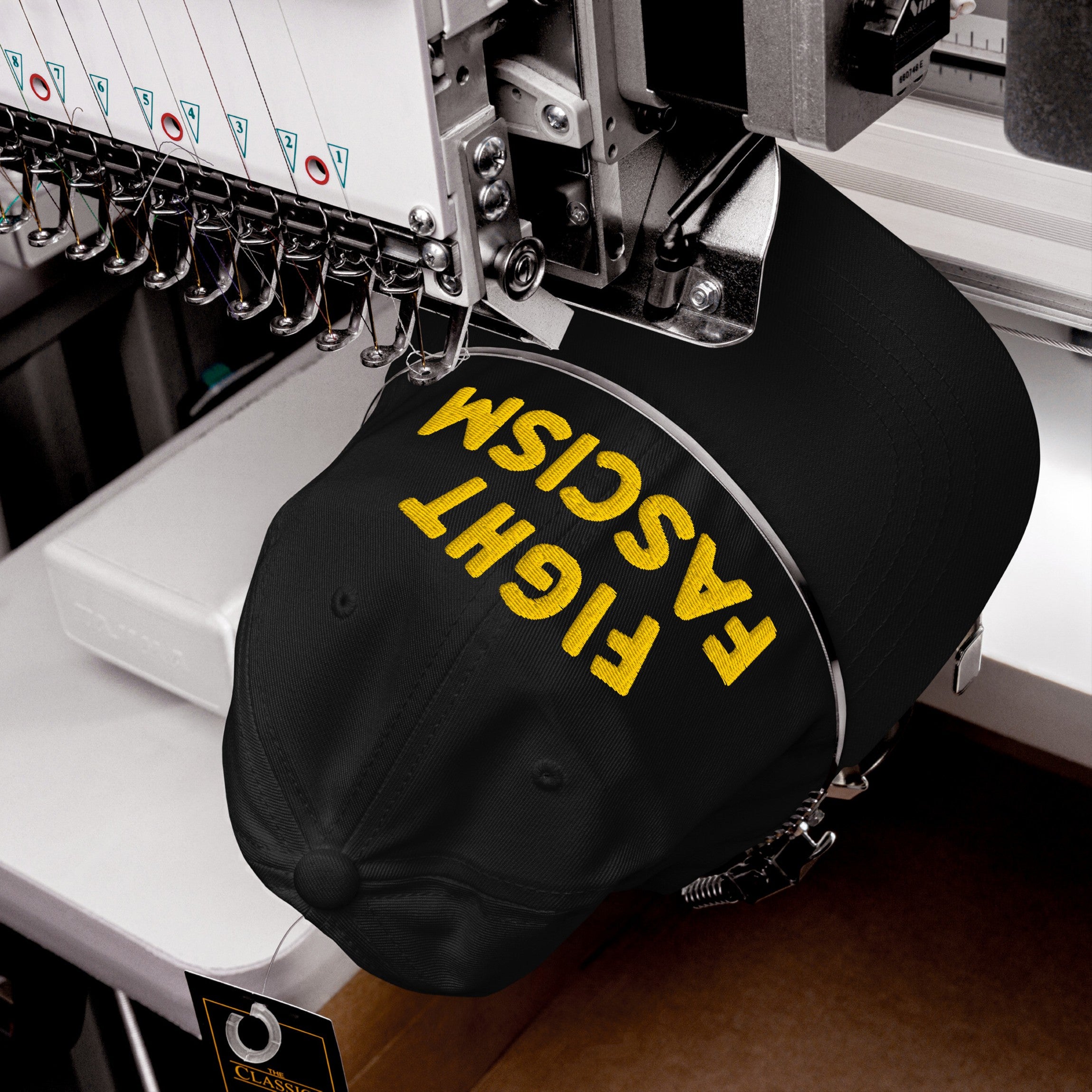 Fight Fascism Hat in embroidery machine, yellow thread stitching bold slogan on black cap, showcasing anti-fascist message for visibility in protests, marches, and daily life.