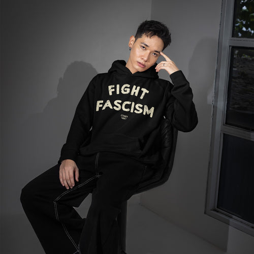 Fight Fascism Hoodie & Sweatshirt on a model seated against a gray-toned backdrop, featuring bold cream letters. The minimalist design reflects a strong anti-fascist message, perfect for activism and daily wear.