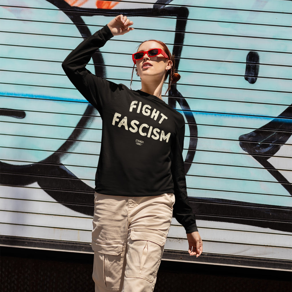 Person wearing Fight Fascism T-shirt with cream distressed lettering, paired with light beige cargo pants and red sunglasses, standing in front of a graffiti-covered shutter, embodying anti-fascist activism.