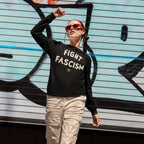 Person wearing Fight Fascism T-shirt with cream distressed lettering, paired with light beige cargo pants and red sunglasses, standing in front of a graffiti-covered shutter, embodying anti-fascist activism.