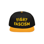 Black snapback-style cap with yellow brim and button, featuring bold yellow FIGHT FASCISM embroidery, embodying protest spiri