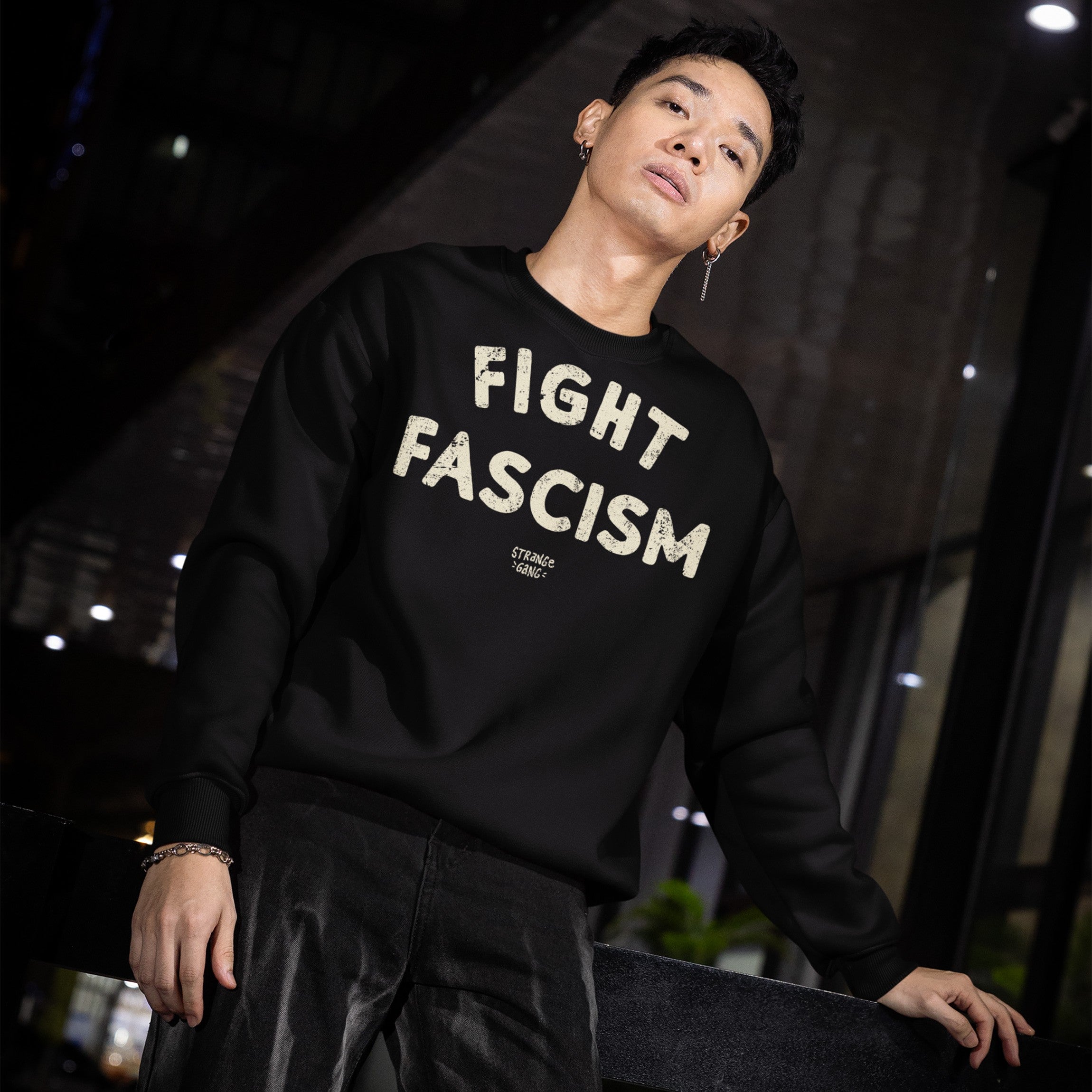 Young man in urban setting wearing Fight Fascism Hoodie & Sweatshirt with prominent cream text, advocating against fascism. Features include short dark hair, earring, bracelet, and dark pants.
