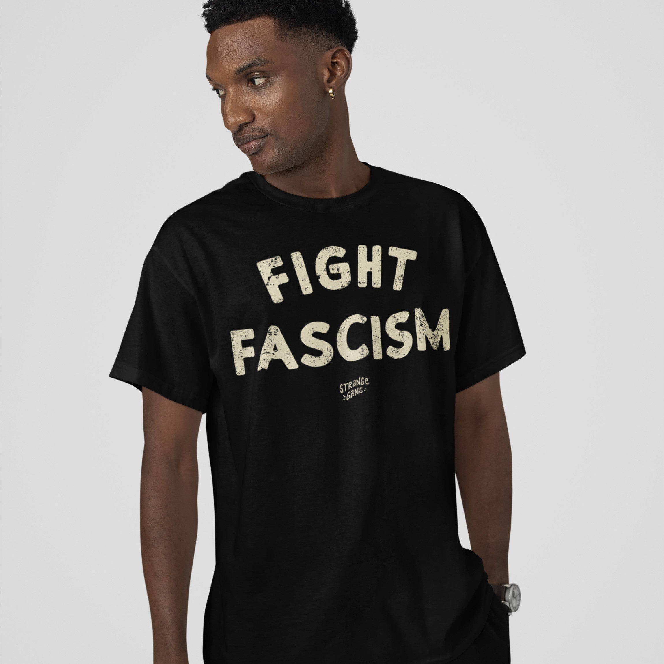 Young man modeling the Fight Fascism T-shirt, featuring large off-white FIGHT FASCISM text and a small logo. He's slightly turned, wearing a hoop earring and wristwatch, against a light-gray background.