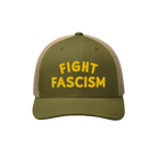 Olive-green trucker-style baseball cap with a tan mesh back, featuring FIGHT FASCISM embroidered in bold yellow letters, desi