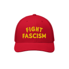 Red mesh-back trucker cap with bold yellow FIGHT FASCISM embroidery on the front, embodying a strong anti-fascist message for