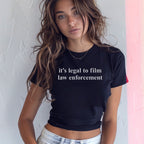 Young woman with wavy brown hair wears a fitted black Film The Police Women's Baby Tee with white text, paired with light-wash denim jeans, standing indoors against a textured wall.