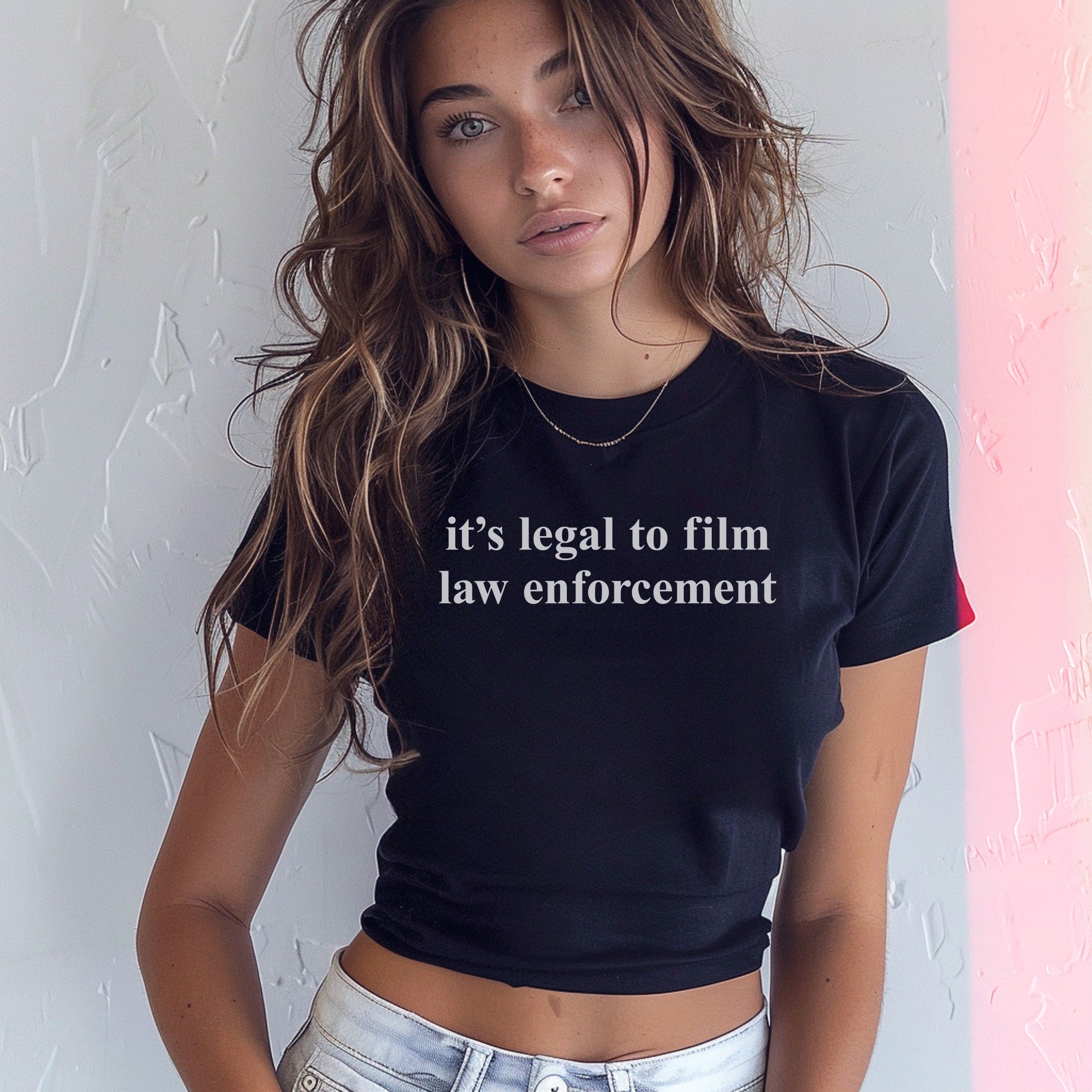 Young woman with wavy brown hair wears a fitted black Film The Police Women's Baby Tee with white text, paired with light-wash denim jeans, standing indoors against a textured wall.