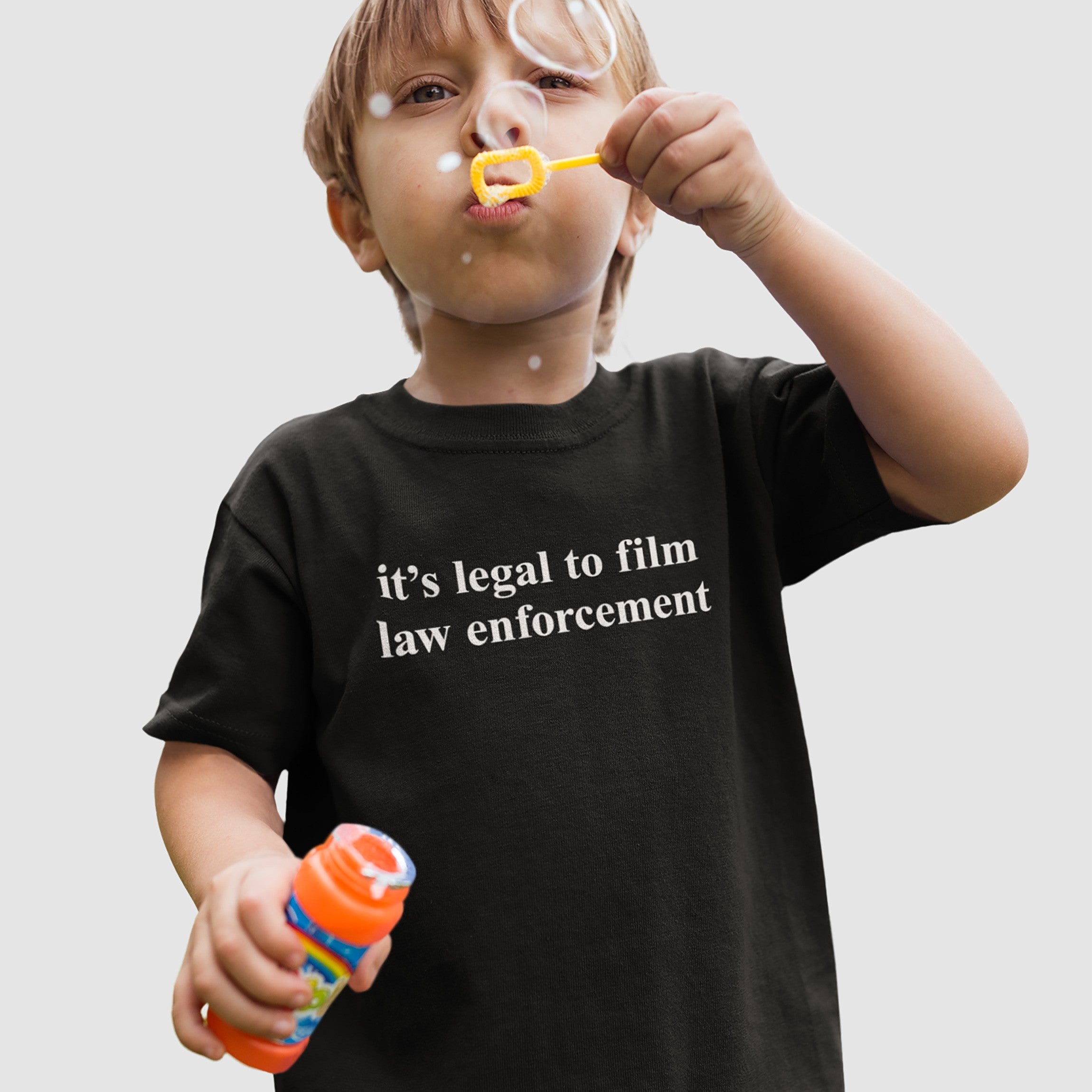 Young child wearing the Film The Police T-shirt, holding a bubble solution and wand, blowing bubbles. The shirt's message, it's legal to film law enforcement, aligns with the store’s advocacy for justice and equality.