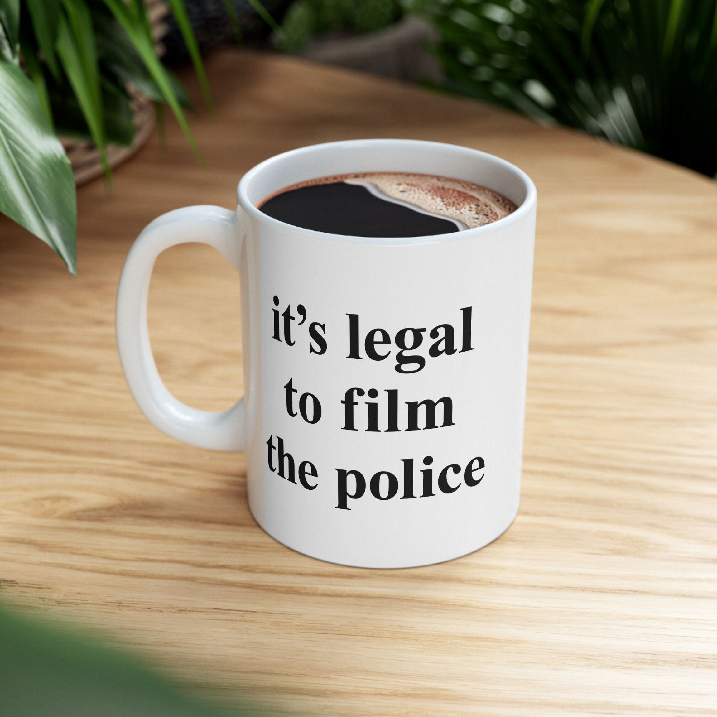 Film The Police Mug: A white ceramic mug on a light wood tabletop filled with black coffee. The mug features the bold statement it's legal to film the police in black type.