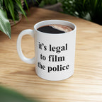 Film The Police Mug: A white ceramic mug on a light wood tabletop filled with black coffee. The mug features the bold statement it's legal to film the police in black type.