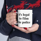 Person holds the Film The Police Mug, a white ceramic cup with bold black lettering. Their red-painted nails and festive sweater add contrast, embodying protest energy. Available in 11 oz. and 15 oz. sizes.