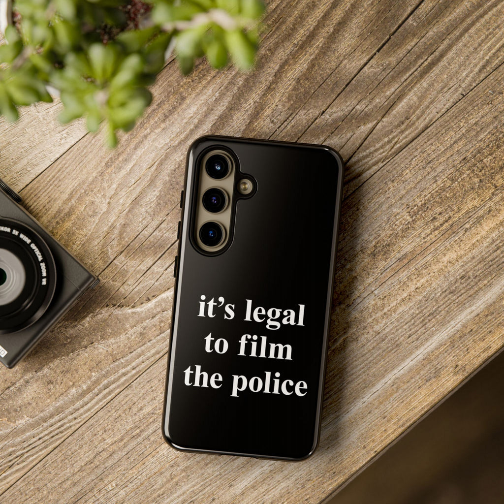 Film The Police Samsung Tough Phone Case on a wooden table, face-down, showcasing its bold slogan. Nearby, a camera lens and blurred foliage complete the setup, emphasizing its activist message.