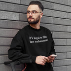 Young man with glasses and a beard wears the Film The Police Hoodie & Sweatshirt, featuring it's legal to film law enforcement text, holding a smartphone, standing beside an electric scooter.