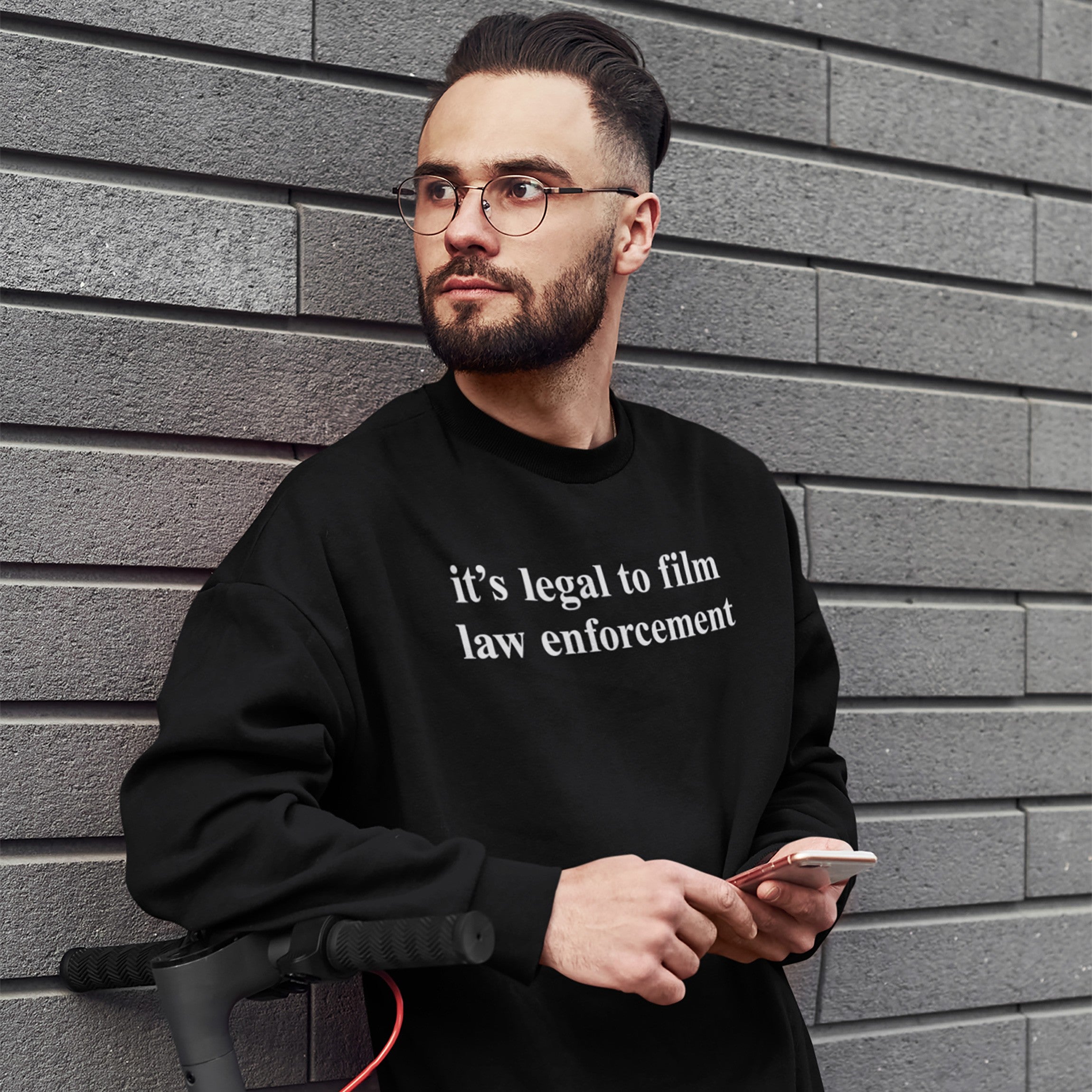 Young man with glasses and a beard wears the Film The Police Hoodie & Sweatshirt, featuring it's legal to film law enforcement text, holding a smartphone, standing beside an electric scooter.